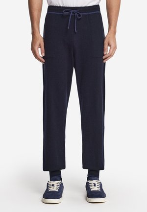 Person trägt eine marineblaue Jogginghose mit Kordelzug, ein weißes T-Shirt, dunkle Socken und marineblaue Sneaker und steht vor weißem Hintergrund.