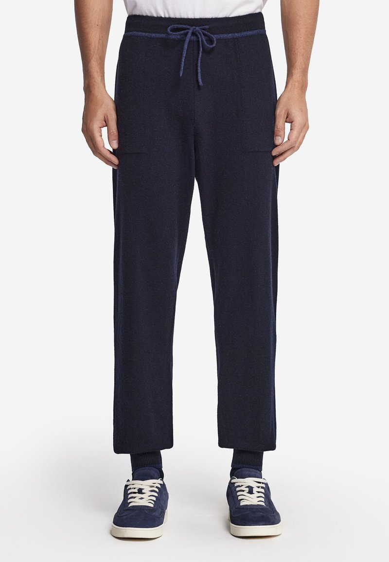 Person trägt eine marineblaue Jogginghose mit Kordelzug, ein weißes T-Shirt, dunkle Socken und marineblaue Sneaker und steht vor weißem Hintergrund.