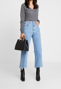 Vrouw in een grijze V-hals trui, lichtblauwe high-waisted jeans met een rafelrand, zwarte enkellaarsjes, een zwarte handtas met bruine handvatten in haar hand.