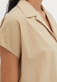 Gros plan d'une personne portant une chemise beige à manches courtes avec col et ouverture en V, montrant une partie du cou et de l'épaule.