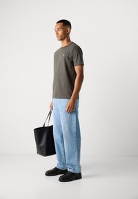 Man in gray t-shirt and light blue jeans holding a black leather tote bag, standing against a plain white background.