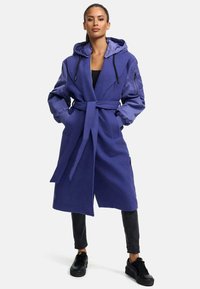 Purple hooded coat with long sleeves, tie waist, and front pockets. Features a contrasting interior lining and ribbed cuffs. Black sneakers worn.