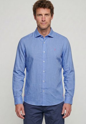 Hombre con camisa azul clara de manga larga abotonada y pantalones oscuros, de pie derecho contra un fondo liso y claro.