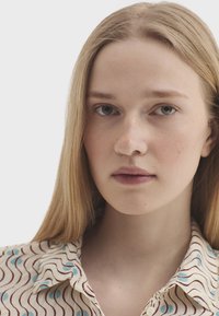 Jeune femme aux longs cheveux blonds portant une chemise à motifs, regardant droit devant avec une expression neutre sur un fond uni.
