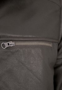 Dunkelgraue Lederjacke mit diagonaler Reißverschlusstasche, metallic-ähnlichem Beschlag und strukturierter Oberfläche. Nahaufnahme des sichtbaren Reißverschlussdetails.
