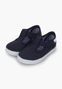 Zapatos de lona azul marino con una correa de velcro y suela de goma blanca. El interior presenta un patrón de cuadros en azul y blanco. Diseño de corte bajo.