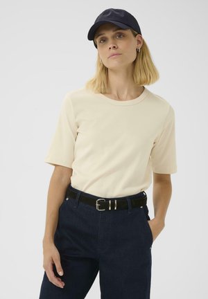 Cremefarbenes T-Shirt mit kurzen Ärmeln, runder Ausschnitt, kombiniert mit dunkelblauen Jeans und einem schwarzen Gürtel mit einer silbernen Schnalle, getragen mit einer dunklen Mütze.
