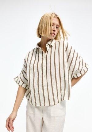 Blonde woman wearing a loose, short-sleeve white shirt with vertical brown stripes and light-colored pants, posing against a white background.