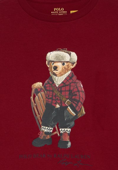 Piros pamut póló, amelyen egy olyan medve illusztráció látható, amely kockás inget, szőrmés sapkát visel és egy szánkót tart. A szöveg tartalmazza: "Polo Bear by Ralph Lauren."