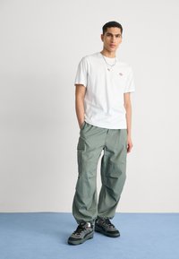 White short-sleeve t-shirt with small logo, green cargo pants with multiple pockets, and dark sneakers with grey and red accents.