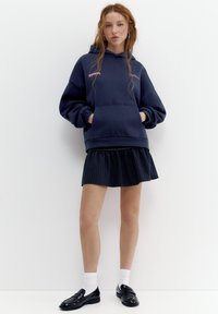 Giovane donna con capelli lunghi che indossa una felpa con cappuccio blu scuro, minigonna a quadri, calzini bianchi e mocassini neri, in piedi contro un muro bianco.