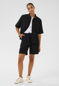 Chemise noire à manches courte et shorts assortis, fabriqués en tissu léger. Dispose d'une fermeture à boutons, de poches et d'une coupe décontractée. Débardeur blanc en dessous.