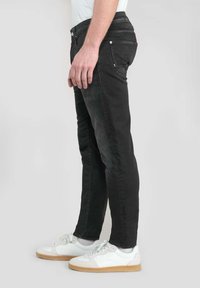 Vaqueros de mezclilla negros con un corte ajustado, que cuentan con un diseño estándar de cinco bolsillos y una superficie texturizada, combinados con zapatillas blancas.
