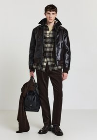 Braune Lederbomberjacke mit schwarzem Schaffellkragen, kariertes Hemd, braune Cordhosen, hält eine schwarze Tasche und einen braunen Schal. Dunkle Schuhe.