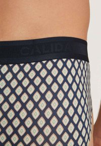 Marineblauwe boxershorts met een geometrisch ruitpatroon in groen en crème. Voorzien van een zwarte elastische band met de merknaam "CALIDA" in reliëf.