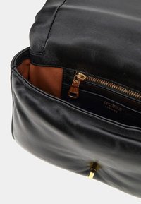 Borsa in pelle nera con una texture liscia, dotata di una chiusura con zip dorata e una foderatura marrone all'interno. Mostra il marchio "Guess" su un'etichetta interna.