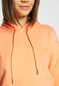 Mikina s kapucňou v žiarivej oranžovej farbe, s jemnou textúrou, dlhými šnúrkami s kovovými koncami a uvoľneným strihom goliera.