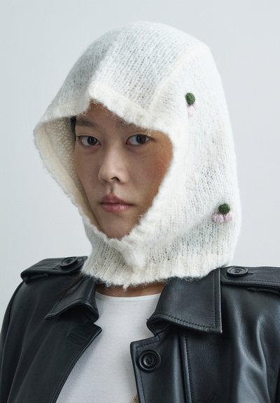 Πλεχτή λευκή κουκούλα με απαλή υφή, με πράσινες και ροζ λεπτομέρειες, φορεμένη πάνω από μια μαύρη δερμάτινη τζάκετ.