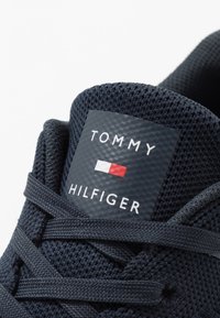 Marine net sneakers med flade snørebånd, udstyret med en sort label, der viser "TOMMY HILFIGER" i hvid og et lille rødt og hvidt logo.