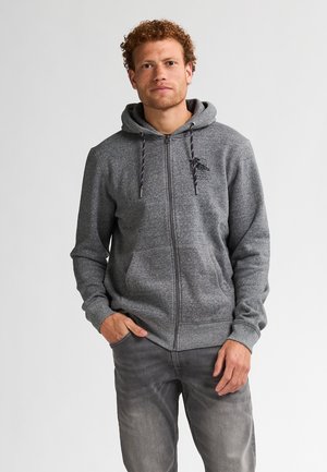 Haut à capuche gris zippé avec cordon de serrage, arborant un logo sur la poitrine, deux poches avant et des poignets et un ourlet côtelés. Porté par-dessus un jean gris clair.