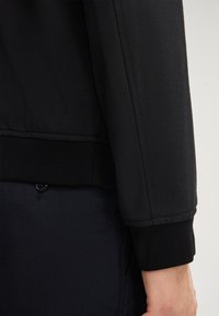 Zwarte langmouwenjas met manchet gedragen over donkere broek met een zichtbare knoopdetail nabij de zak, weergegeven op een staande persoon.