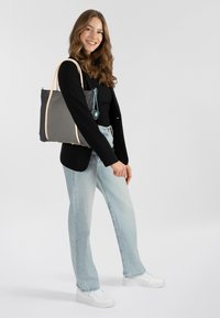 Grijze tote bag met crèmekleurige accenten, textuur oppervlak en blauwe hardware, vastgehouden door een persoon in een zwarte top en lichte jeans tegen een effen achtergrond.