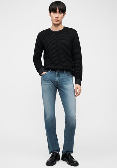 Άνδρας που φοράει υφασμένο μαύρο πουλόβερ με μακριά μανίκια, μπλε τζιν με μαύρη ζώνη και μαύρα παπούτσια, στέκεται με το ένα χέρι στην τσέπη.