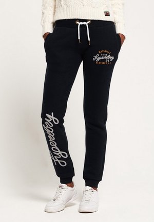 Black jogger pants with white drawstring, "Superdry" script on left leg, and small "Maysville Kentucky" logo on right hip, worn with white sneakers.