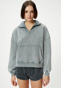Sudadera gris de gran tamaño hecha de un material suave; cuenta con un cuello de media cremallera, bolsillo frontal y puños de canalé. Combinada con pantalones cortos oscuros.