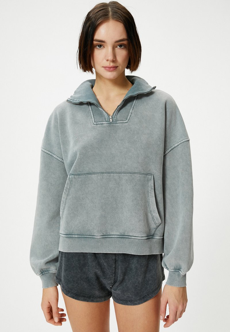 Sudadera gris de gran tamaño hecha de un material suave; cuenta con un cuello de media cremallera, bolsillo frontal y puños de canalé. Combinada con pantalones cortos oscuros.