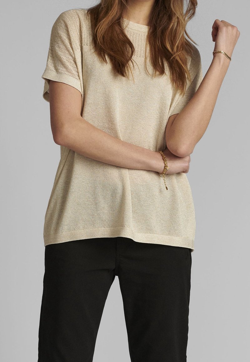 Femme portant un haut en maille beige à manches courtes et un pantalon noir, avec des bracelets dorés aux deux poignets, debout devant un fond uni.