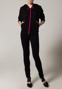Felpa nera con zip e cordini rosa, abbinata a pantaloni neri aderenti. La felpa presenta una tasca frontale e maniche corte. Scarpe aperte nere.