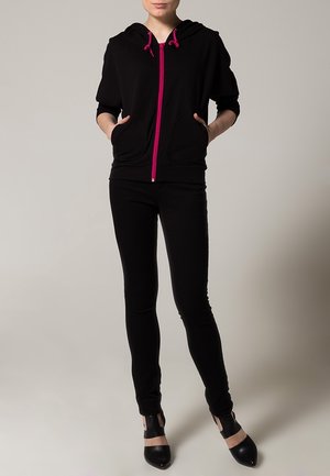 Czarna bluza z kapturem z różowym zapięciem i sznurkami, w połączeniu z dopasowanymi czarnymi spodniami. Bluza ma kieszeń z przodu i krótkie rękawy. Czarne buty z odkrytymi palcami.