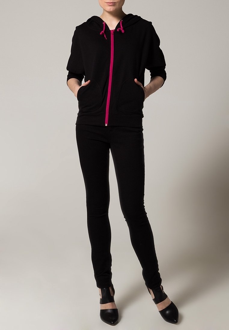 Felpa nera con zip e cordini rosa, abbinata a pantaloni neri aderenti. La felpa presenta una tasca frontale e maniche corte. Scarpe aperte nere.