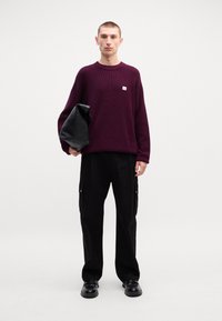 Maroon knit sweater with small logo, paired with loose black cargo pants featuring side pockets. Black bag held in right hand, shiny shoes.