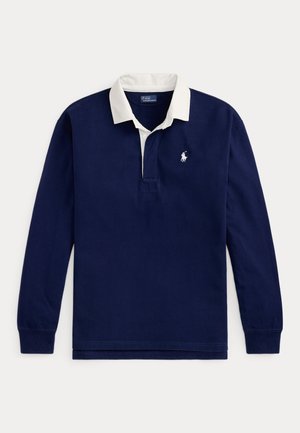 Femme debout portant un polo bleu marine à manches longues, un pull beige noué à la taille, un pantalon vert olive et des baskets blanches sur fond neutre.