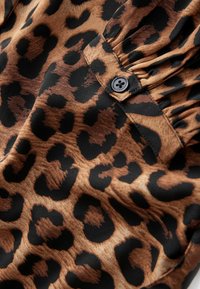 Tissu à imprimé léopard avec des motifs marron et noir ; comprend un détail bouton et des fronces, avec une texture lisse et douce.