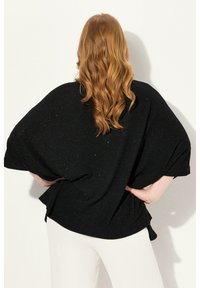 Haut noir surdimensionné avec une coupe ample et des manches chauve-souris, présentant une texture scintillante subtile sur un fond clair.
