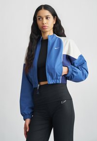 Veste courte Nike bleu et blanc, tissu léger, poignets élastiques, fermeture éclair complète et logo brodé ; associée à un legging noir.