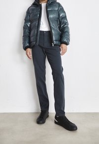 Chaqueta acolchada de color teal oscuro con cierre de cremallera, usada sobre una camiseta blanca. Combinada con pantalones oscuros y zapatillas negras con acentos blancos.