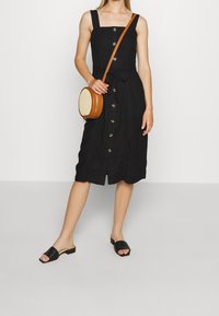 Svart midiklänning med fyrkantig halsringning, framtida knappar och knytmidja. Matchad med svarta slidesandaler och en rund beige axelväska.