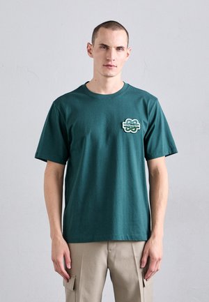 Mørkegrøn bomulds t-shirt med et broderet logo foran i en hvid og grøn kløverform. Basic crew neck og korte ærmer.