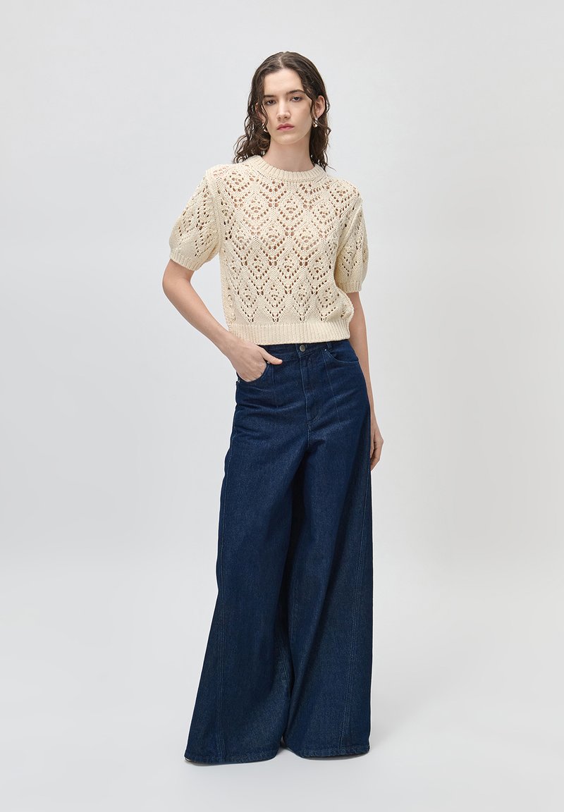 Femme portant un pull court à manches courtes en maille crème et un jean taille haute bleu marine à jambes larges, debout devant un fond uni.