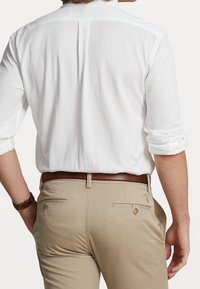 Vit skjorta med uppvuxna ärmar, matchad med beige byxor och ett brunt läderbälte. Bakifrån framhävs detaljer som sömmar och fickor.