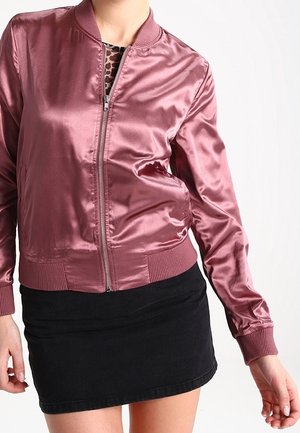 Nainen, jolla on päällään kiiltävä mauvenvärinen vetoketjullinen bomber-takki mustan minihameen päällä, seisoo yksinkertaisen valkoisen taustan edessä.