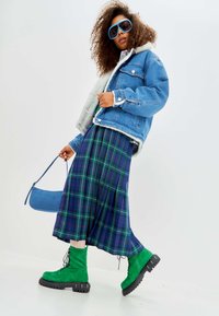 Jeansjack met schapenwolvoering, groen geruite plissérok en groene veterschoenen met een chunky zool, gecombineerd met een blauwe handtas.