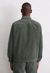 Zielona kurtka ze sztruksu z pionowymi paskami, z kołnierzem i mankietami zapinanymi na guziki, wykonana z teksturowanej tkaniny, widziana od tyłu.