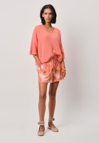 Koraalroze fuzzy brei V-hals trui, gecombineerd met roze tie-dye shorts. De look wordt afgemaakt met beige flats en enkelbandjes.