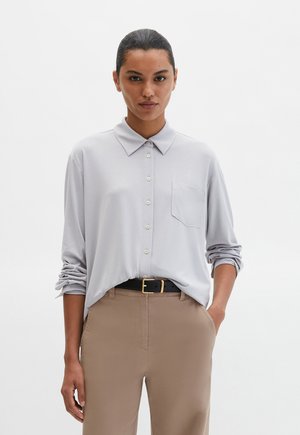 Blouse gris clair à boutons avec manches longues et une poche poitrine, associée à un pantalon beige et une ceinture noire avec une boucle dorée.