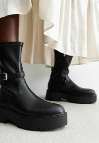 Svarta läderankelboots med en kraftig platåsula, med spänne detaljer och rund tå, placerade mot en ljus bakgrund.